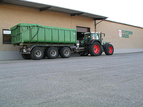 Ein Traktor mit einem langen, grünen Anhänger steht vor einer hellbraunen Hausfassade mit der Aufschrift P H Petersen Saatzucht Lundsgaard.