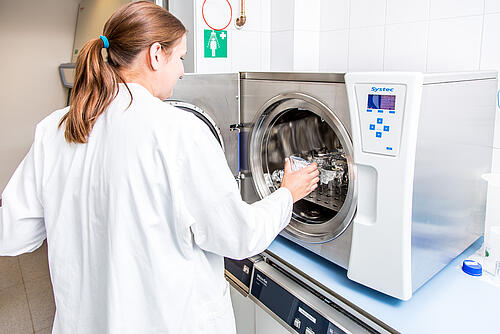 Eine Frau in weißem Kittel hält einen mit Alufolie abgedeckten Glasbehälter in der Hand. Sie steht an der offenen Tür einer weißen Maschine mit der Aufschrift Systec.